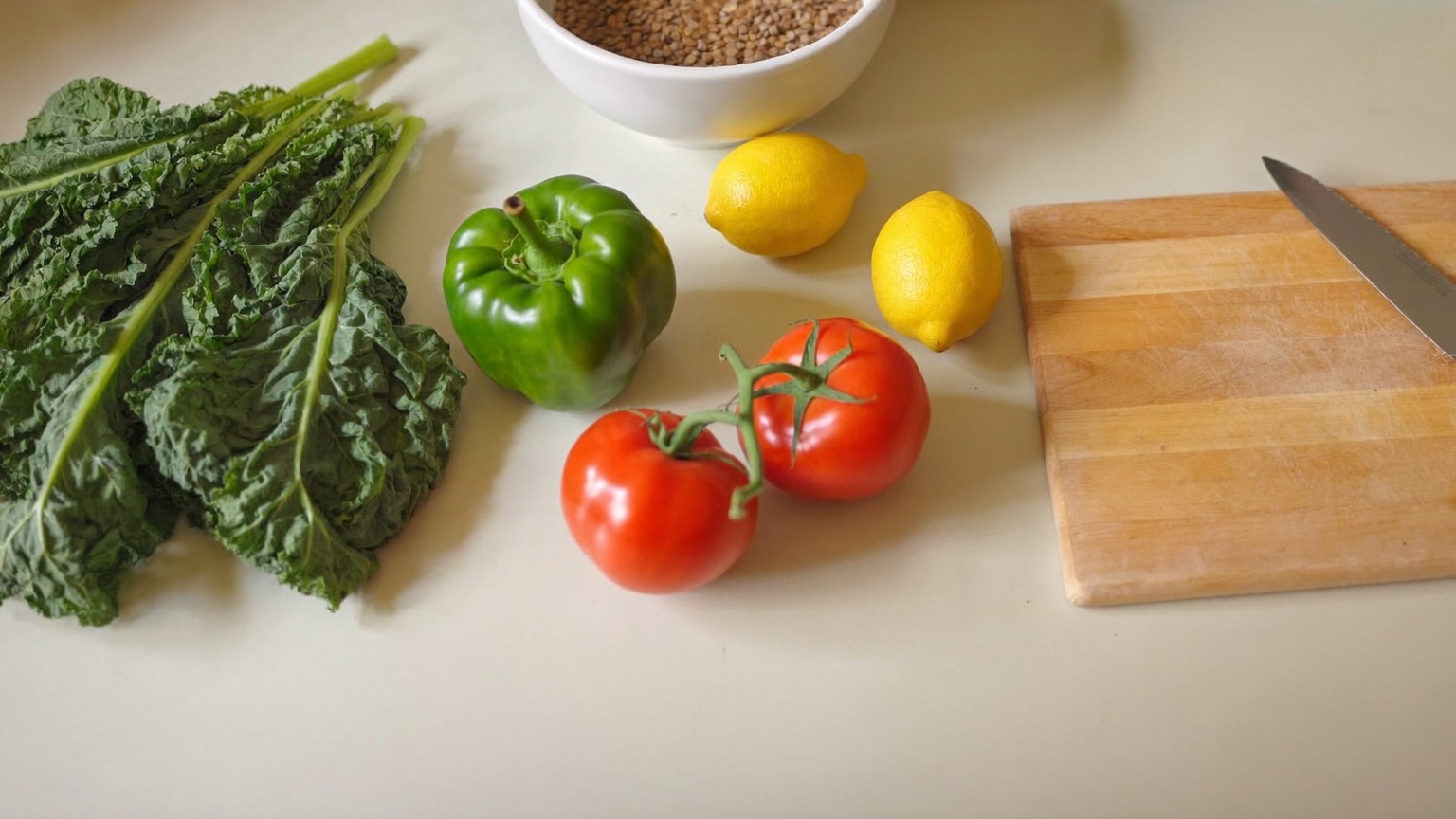 An overhead view of fresh kale, a green bell pepper, vine tomatoes, two lemons, a white bowl of dried grains, and a wooden cutting board with a knife on a pale kitchen counter in warm morning light