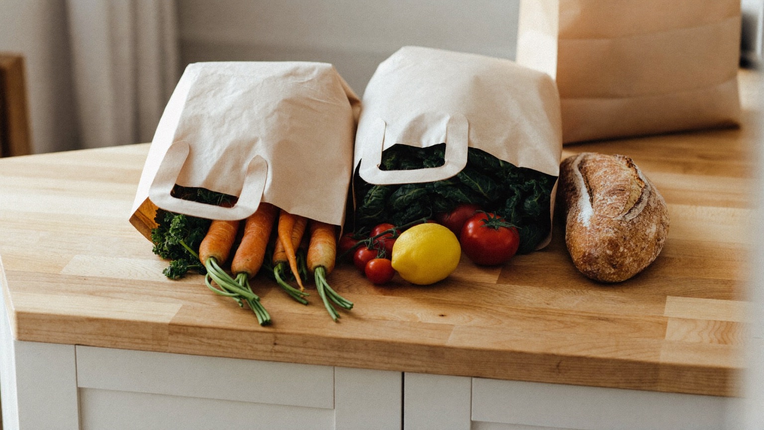 Two plain brown paper grocery bags standing on a light-wood kitchen counter in soft daylight, fresh produce at their open tops — carrots with green stems, a bunch of dark kale, red tomatoes, and a yellow lemon — with a rustic loaf of bread beside them