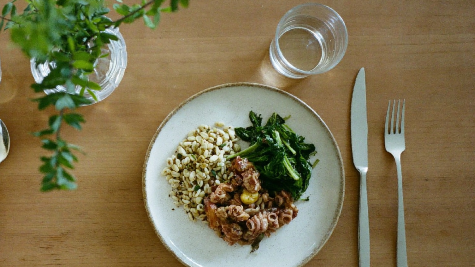 An overhead photo of a home-cooked dinner of grain, sauteed greens, and pasta on a cream stoneware plate beside a water glass, fork and knife, and a small vase of greenery on a wooden table