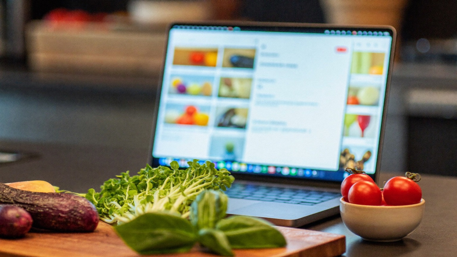 A laptop open on a kitchen counter behind a cutting board of fresh vegetables
