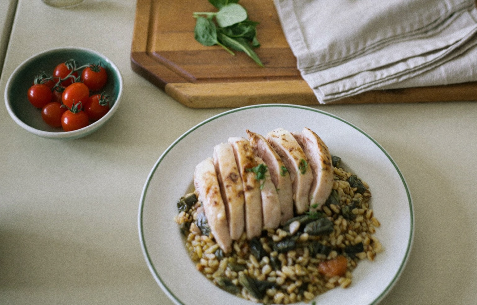 An overhead view of a home-cooked weeknight dinner on a warm off-white table — roasted chicken with grains and greens, a bowl of cherry tomatoes, and fresh herbs on a wooden board in soft natural light