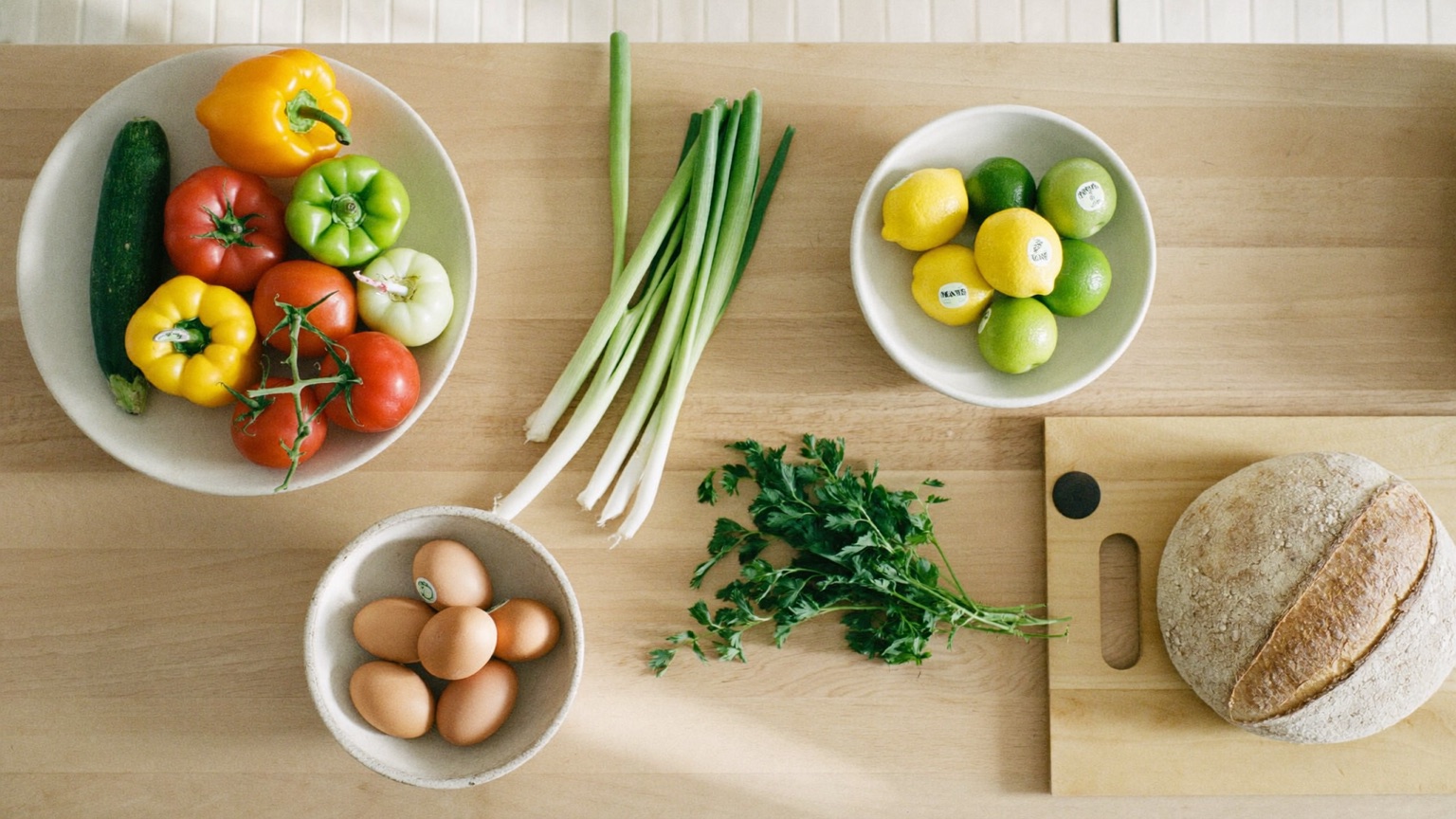 Fresh whole-food groceries in separate bowls on a light-wood counter — bell peppers and tomatoes, scallions, citrus fruit, brown eggs, parsley, and a round bread loaf on a cutting board