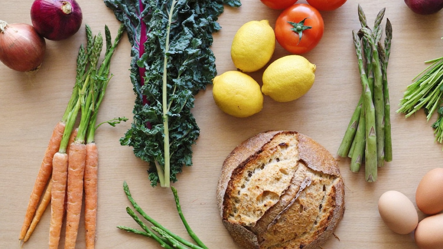 An overhead spread of loose fresh produce, eggs, and a sourdough loaf on a wood kitchen counter