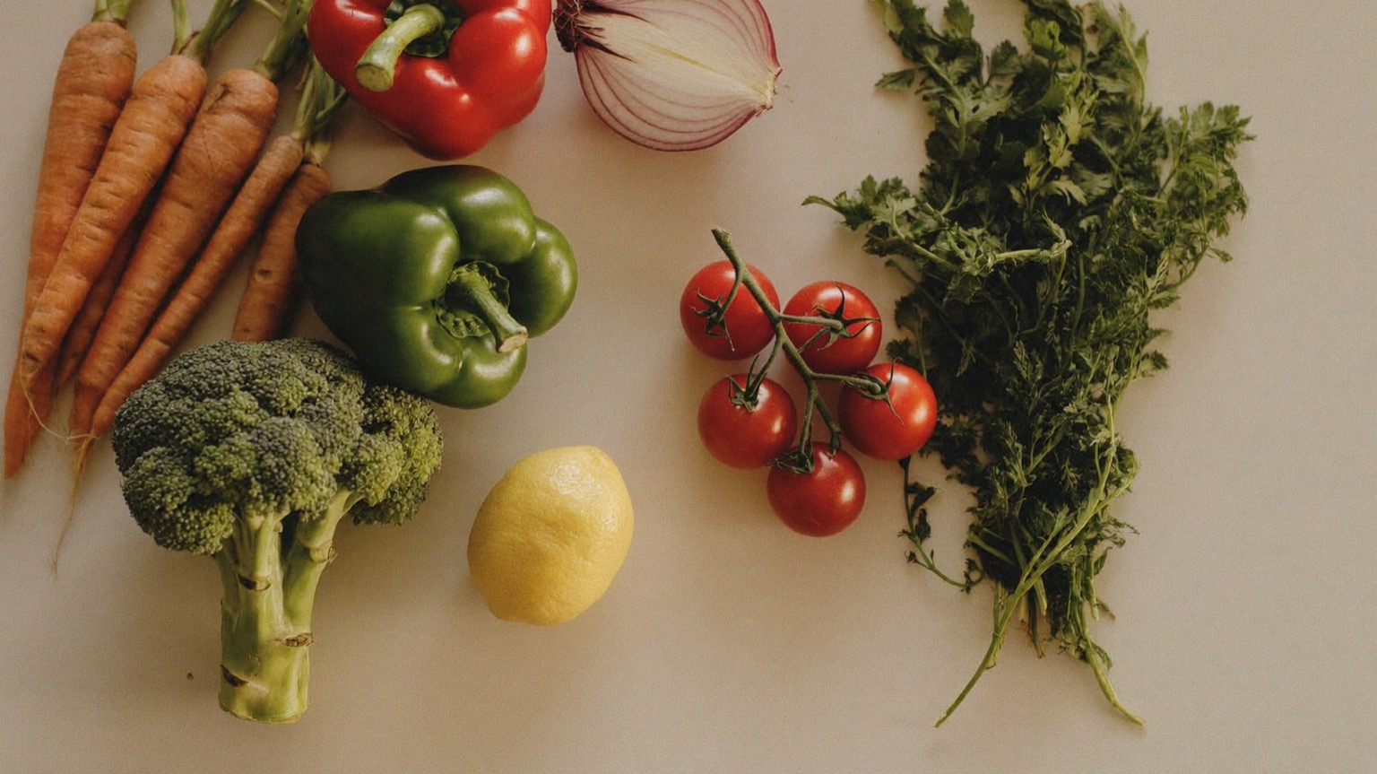 Fresh whole vegetables — carrots, red and green bell peppers, broccoli, a halved red onion, cherry tomatoes on the vine, herbs, and a lemon — arranged on an off-white kitchen counter in warm natural light