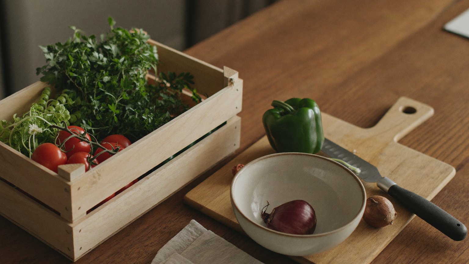 A wooden crate of fresh herbs and vine tomatoes beside a cutting board holding a green bell pepper, a bowl with a red onion, and a knife on a wooden kitchen table in warm light