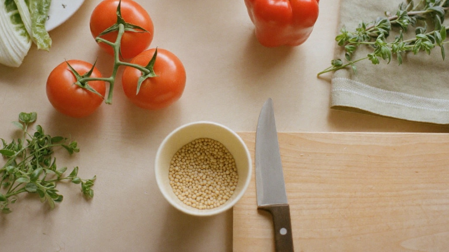 An overhead view of a warm kitchen counter with vine tomatoes, leafy greens, a red bell pepper, fresh herbs, a bowl of grains, a wooden cutting board with a chef's knife and a sage-green linen towel in soft morning light