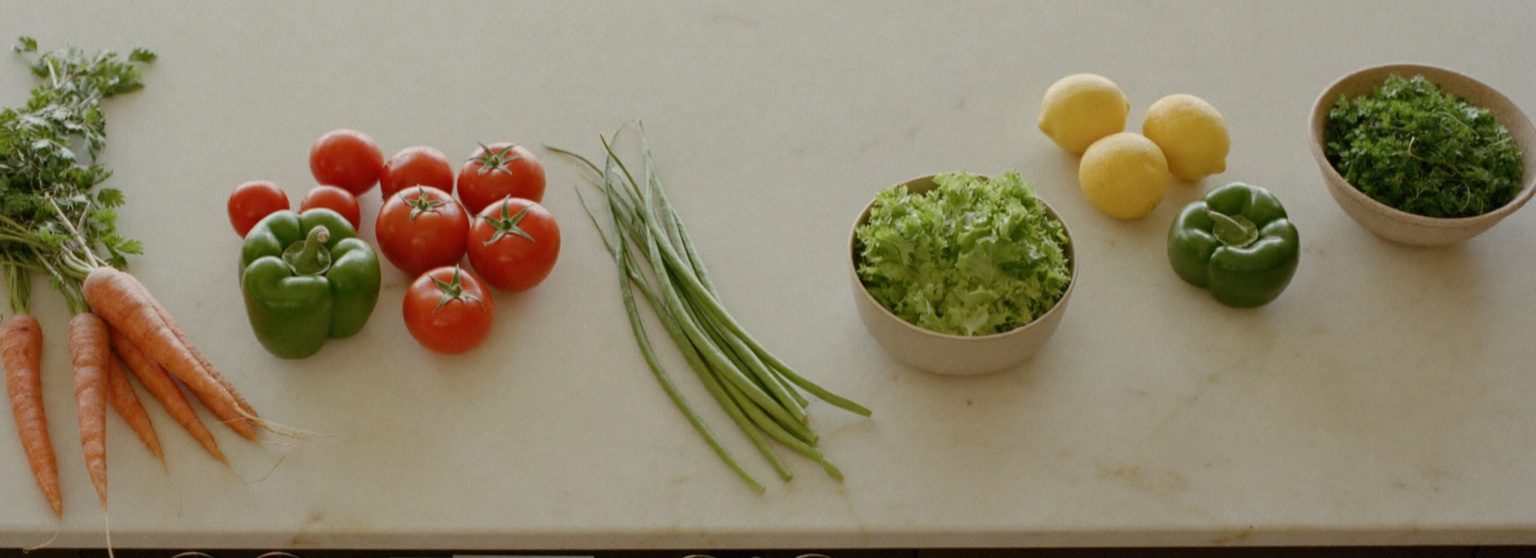 Fresh vegetables and fruit — carrots, a green bell pepper, ripe tomatoes, green beans, a bowl of leafy lettuce, lemons, and a bowl of parsley — laid out in a row on an off-white marble kitchen counter in soft natural light