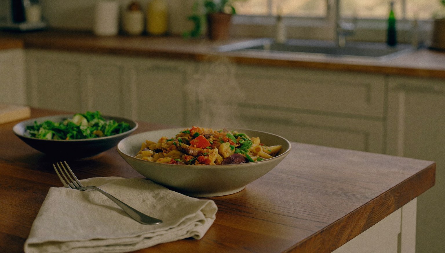 A home-cooked pasta dinner with a side salad, freshly plated on a warm wooden kitchen counter in soft evening window light